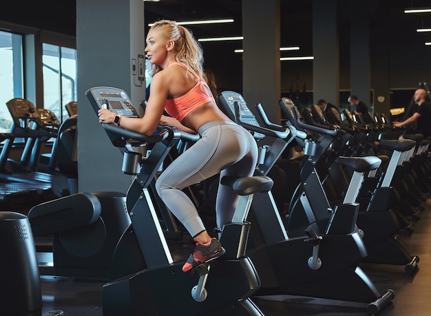 Bezpłatne zdjęcie sportive blond kobieta ćwiczy na rowerze stacjonarnym w nowoczesnym centrum fitness. styl życia fitness w klubie sportowym.