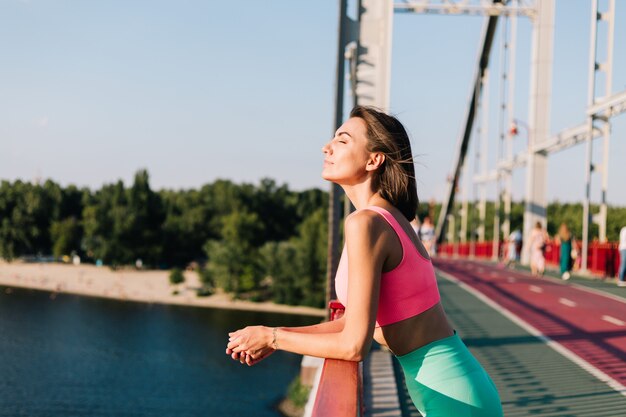 Sportowa kobieta w dopasowanym stroju sportowym o zachodzie słońca na nowoczesnym moście z widokiem na rzekę rozejrzyj się, ciesząc się letnią pogodą