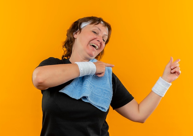 Bezpłatne zdjęcie sportowa kobieta w średnim wieku w czarnej koszulce z opaską i ręcznikiem na ramieniu, uśmiechnięta wesoło, wskazująca palcami w bok, stojąca nad pomarańczową ścianą
