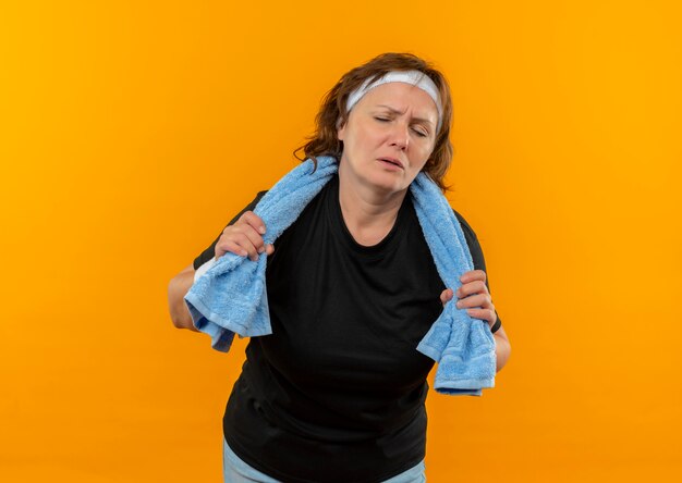 Sportowa kobieta w średnim wieku w czarnej koszulce z opaską i ręcznikiem na ramieniu wyglądająca na zmęczoną i wyczerpaną po treningu, stojąca nad pomarańczową ścianą