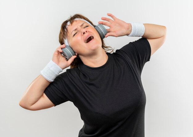 Sportowa kobieta w średnim wieku w czarnej koszulce z pałąkiem na głowę i słuchawkami szalona szczęśliwa słuchająca muzyki stojącej na białej ścianie