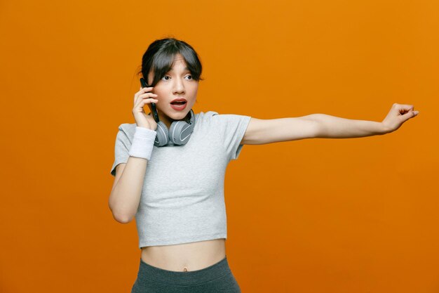 Sportowa piękna kobieta w odzieży sportowej ze słuchawkami rozmawiająca przez telefon komórkowy, wyglądająca na zaskoczoną stojącą na pomarańczowym tle