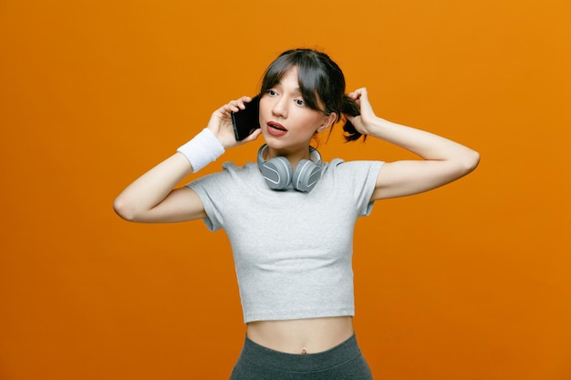 Sportowa piękna kobieta w odzieży sportowej ze słuchawkami rozmawiająca przez telefon komórkowy, wyglądająca pewnie i zaintrygowana, stojąc na pomarańczowym tle