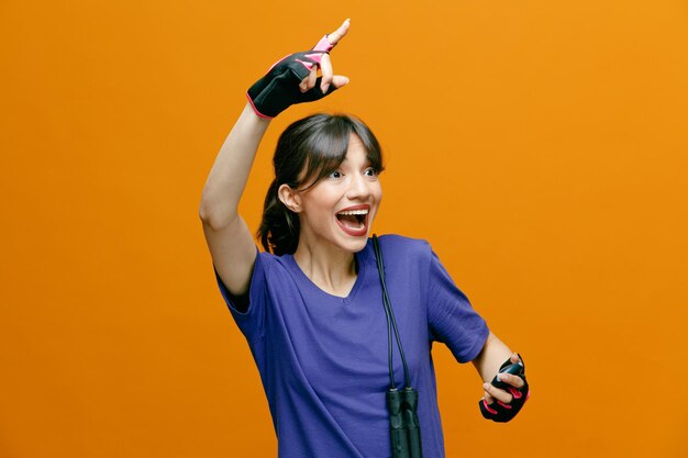 Sportowa piękna kobieta w stroju sportowym w rękawiczkach ze skakanką na ramieniu, trzymająca stoper patrząc na bok, szczęśliwa i podekscytowana, wskazując palcem wskazującym w górę, stojąc na pomarańczowym tle