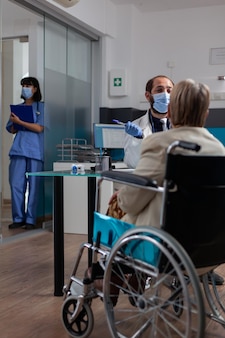 Spotkanie lekarza ze starym pacjentem na wózku inwalidzkim podczas badania kontrolnego podczas pandemii covid 19. specjalista udzielający wsparcia i porad dla rencisty z niepełnosprawnością ruchową w gabinecie lekarskim.