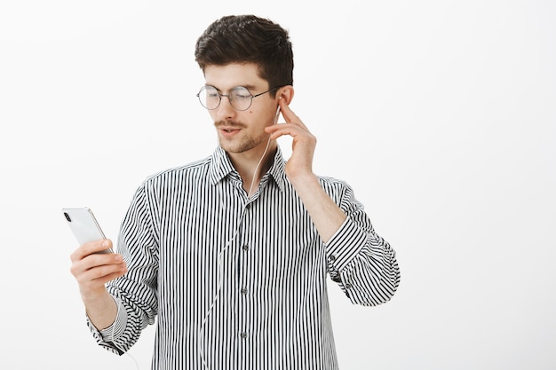 Sprawdzam świetny Utwór Ulubionej Piosenkarki. Portret Skupionego Przystojnego, Zdumionego Mężczyzny Z Wąsami, Trzymającego Smartfon, Wybierającego Piosenkę Podczas Słuchania Muzyki W Słuchawkach Na Szarej ścianie