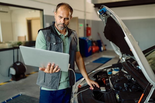 Średni dorosły mechanik prowadzący diagnostykę samochodową na laptopie w warsztacie samochodowym