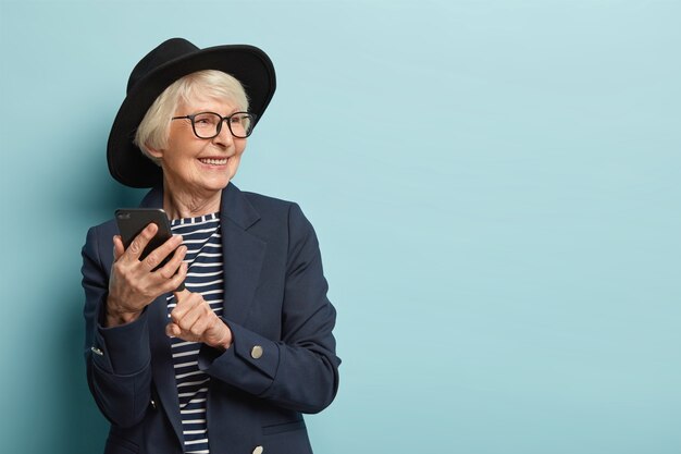 Stara atrakcyjna kobieta pisze SMS-y przez komórkę, korzysta z nowej aplikacji online, ma pozytywny wyraz twarzy, odwraca wzrok, nosi stylowe nakrycie głowy i płaszcz, stoi nad niebieską ścianą, wolne miejsce