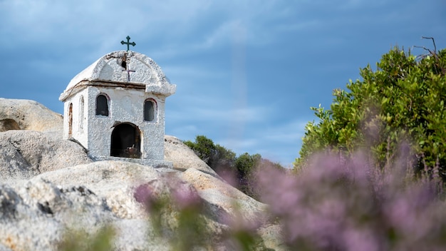 Stara i mała kapliczka położona na skałach w pobliżu wybrzeża Morza Egejskiego, dookoła krzaki, zachmurzone niebo, Grecja