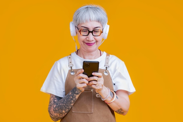 Stara starsza azjatycka kobieta nosić fartuch ręcznie wybierając listę odtwarzania muzyki ze smartfona zrelaksować się dorywczo styl życiaasia tatuaż kobieta nosić słuchawki słuchać muzyki beztroski izolować żółte tło studio strzał