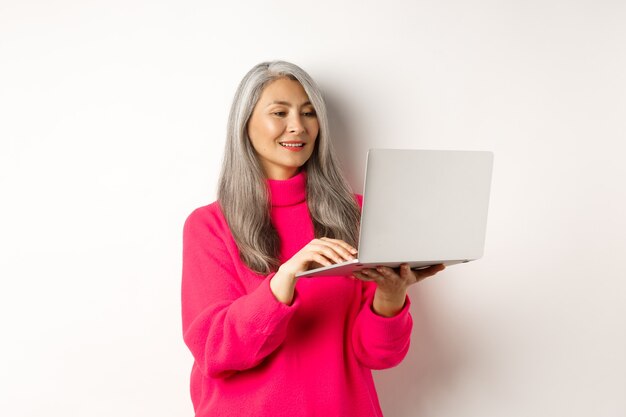 Starsza azjatycka kobieta pracuje jako freelancer używając laptopa i uśmiechając się stojąc na białym tle