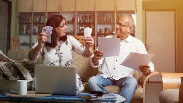 Starsza indyjska para azjatycka księgowa, robi finanse domowe i sprawdza rachunki za pomocą laptopa, kalkulatora i pieniędzy również ze skarbonki, siedząc na kanapie lub stole w domu