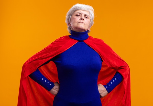 Bezpłatne zdjęcie starsza kobieta superbohaterka w czerwonej pelerynie, patrząca na kamerę z pewnym siebie wyrazem twarzy z rękami na biodrze, stojąca nad pomarańczą