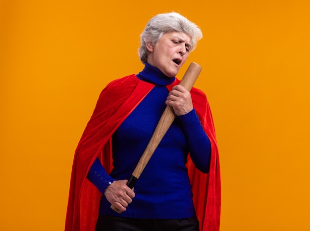 Bezpłatne zdjęcie starsza kobieta superbohaterka w czerwonej pelerynie trzyma kij bejsbolowy, używając go jako mikrofonu