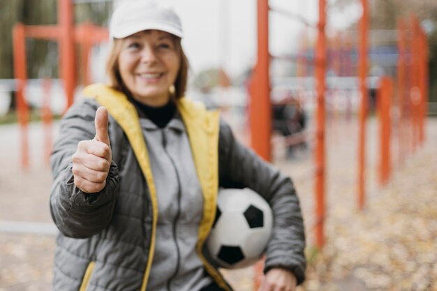Starsza kobieta trzyma piłkę nożną i daje kciuki do góry podczas ćwiczeń