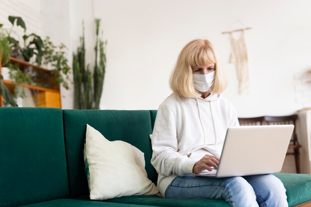 Starsza kobieta używa laptopa w domu z maską medyczną