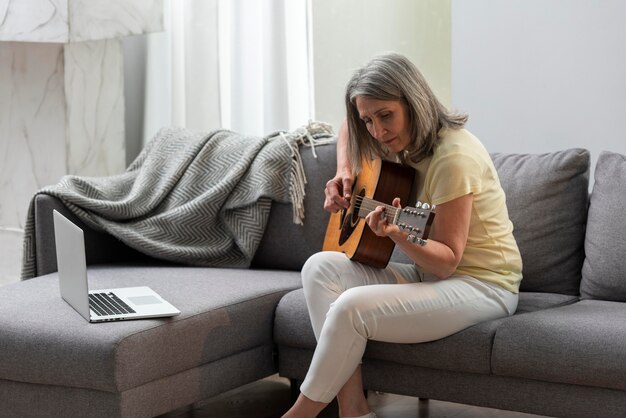 Starsza kobieta w domu na kanapie za pomocą laptopa do lekcji gry na gitarze