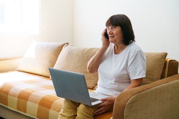 Starsza kobieta z laptopem siedząca na kanapie rozmawiająca z kimś przez telefon nowe technologie są ...