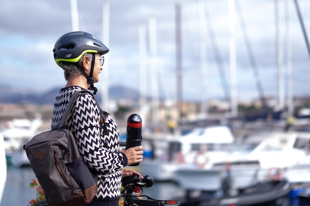 Starsza rowerzystka w plecaku i kasku, trzymająca butelkę z wodą, patrząca na łodzie w porcie