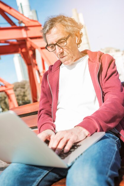 Starszego mężczyzna obsiadanie na ławce pisać na maszynie na laptopie przy outdoors