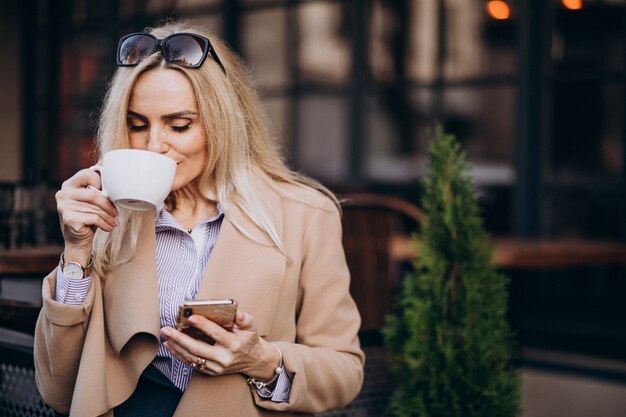 Starszy bizneswoman pije kawę na zewnątrz kawiarni i rozmawia przez telefon
