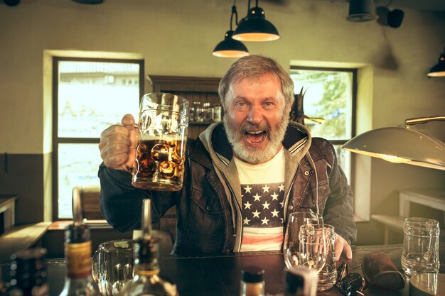 Starszy brodaty mężczyzna pije alkohol w pubie i ogląda program sportowy w telewizji. Cieszę się moją ulubioną kawą i piwem. Mężczyzna z kuflem piwa siedzi przy stole.