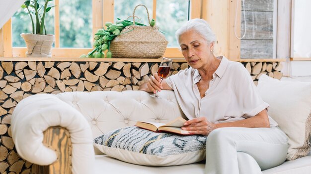 Starszy kobiety obsiadanie na kanapie trzyma wina szkła czytelniczą książkę
