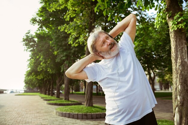 Starszy mężczyzna jako biegacz na ulicy miasta. Kaukaski mężczyzna model jogging i trening cardio w letni poranek. Wykonywanie ćwiczeń rozciągających w pobliżu łąki. Zdrowy styl życia, sport, koncepcja aktywności.