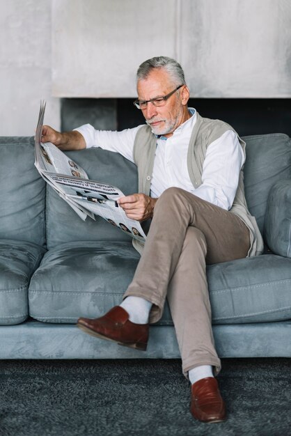 Starszy mężczyzna siedzi na wygodnej kanapie ze skrzyżowanymi nogami czytanie gazety