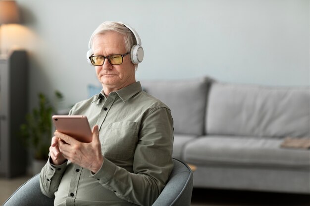 Starszy mężczyzna słucha muzyki ze słuchawkami i tabletem, siedząc na kanapie w salonie