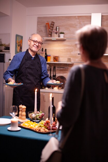 Starszy mężczyzna służy żonie podczas celebrowania ich związku ze smacznym i winem. Starsza para rozmawia, siedzi przy stole w kuchni, jedząc posiłek, świętując swoją rocznicę.
