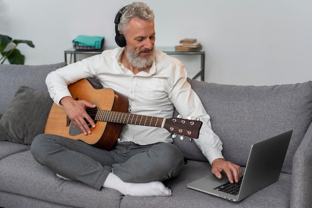 Starszy mężczyzna w domu na kanapie za pomocą laptopa do nauki lekcji gry na gitarze
