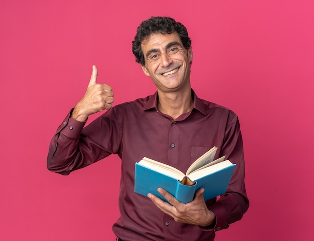 Starszy Mężczyzna W Fioletowej Koszuli Trzyma Otwartą Książkę, Patrząc Na Kamerę, Uśmiechając Się Radośnie Pokazując Kciuk Do Góry Stojący Nad Różowym Tłem