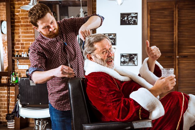 Starszy mężczyzna w stroju Świętego Mikołaja goli swojego osobistego mistrza w salonie fryzjerskim przed świętami Bożego Narodzenia