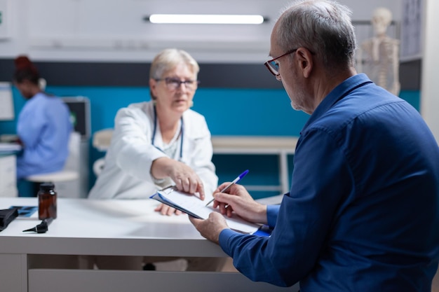 Starszy pacjent podpisujący dokumenty dotyczące wizyty u lekarza i systemu opieki zdrowotnej w gabinecie. chory człowiek robi podpis na plikach schowka, aby otrzymać lek na receptę podczas wizyty kontrolnej.