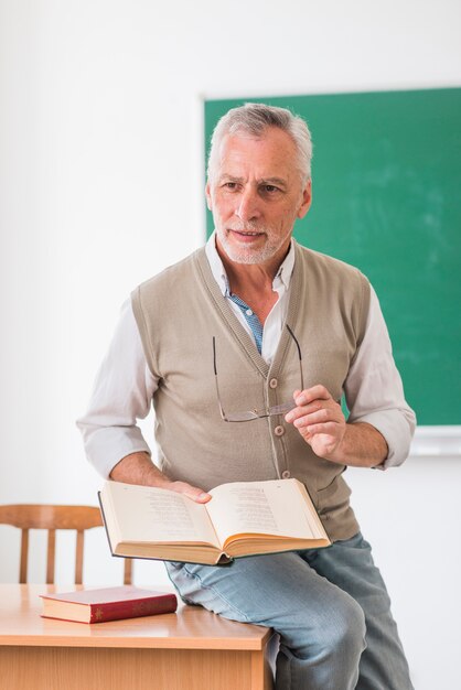 Starszy profesor siedzi na biurku z książką w klasie