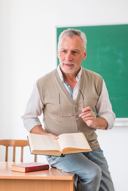 Starszy profesor siedzi na biurku z książką w klasie