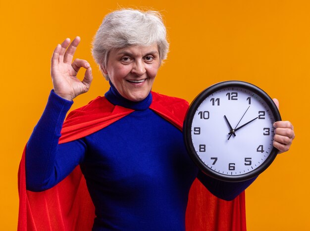 Starszy superbohaterka nosząca czerwoną pelerynę trzymająca zegar patrząc na kamerę szczęśliwa i wesoła robi ok znak stojący na pomarańczowym tle
