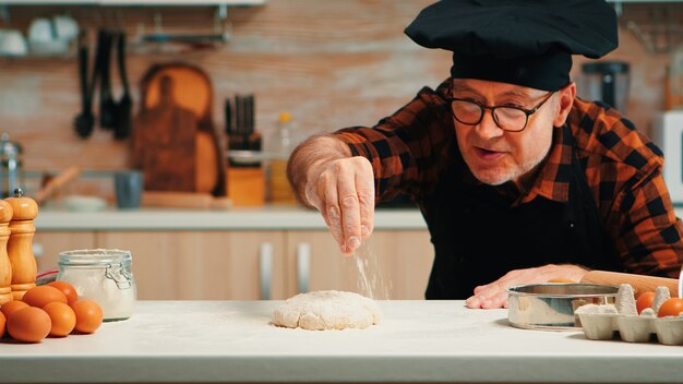 Starszy szef kuchni z bonete co do pieczenia ciasta. Emerytowany starszy piekarz z fartuchem, posypywanie mundurem kuchennym, przesiewanie, rozprowadzanie składników rew z ręcznym pieczeniem domowej pizzy i chleba.