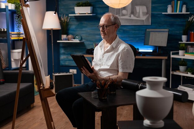 Starszy twórca sztuki przeglądający cyfrowy tablet, szukający inspiracji w internetowej galerii sztuki w domowym studiu ze sztalugą. Starszy artysta korzystający z cyfrowego urządzenia do doskonalenia techniki rysunkowej w warsztacie.