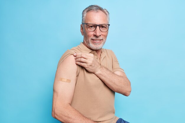 Staruszek z siwymi włosami pokazuje zaszczepione ramię motywuje do zaszczepienia się przeciwko koronawirusowi, aby zatrzymać epidemię, troszczy się o zdrowie w jego wieku, nosi okulary w brązowym t-shirtie, odizolowane na niebieskiej ścianie