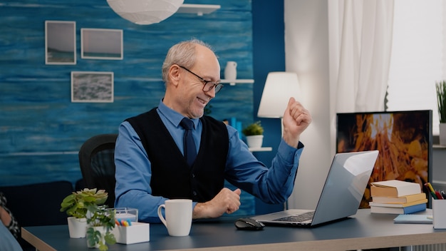 Stary freelancer odbiera dobre wieści na laptopie pracującym w domu, siedząc w salonie. Entuzjastyczny starszy pracownik korzystający z nowoczesnej technologii, czytający, piszący, szukający, gdy starsza żona siedzi na kanapie