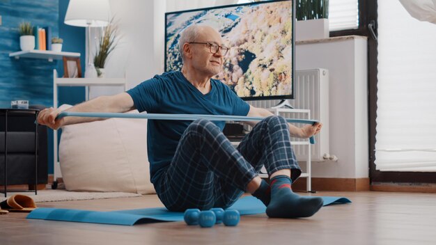 Stary mężczyzna używający opaski do rozciągania i ciągnięcia, siedzący na macie do jogi. Trening seniora z elastycznym pasem do ćwiczeń mięśni ramion w domu. Starszy dorosły wykonujący aktywność fizyczną