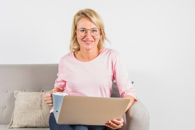 Bezpłatne zdjęcie starzejąca się uśmiechnięta kobieta w różanej bluzce z laptopem i filiżanką na kanapie