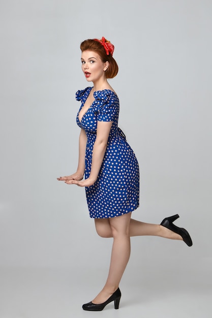 Bezpłatne zdjęcie stereotypowy obraz emocjonalnej, zabawnej pin up girl w stylowym stroju vintage ze zdumionym wyrazem twarzy, otwierającymi usta, wykonującą zalotny gest i podnoszącą nogę do tyłu
