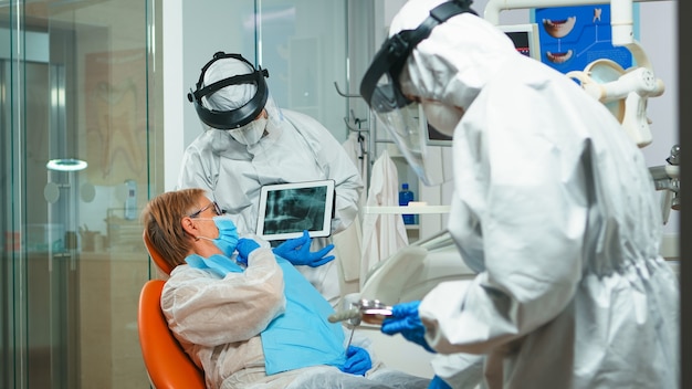 Stomatolog w kombinezonie ochronnym przeglądający prześwietlenie zęba ze starszym pacjentem wyjaśniającym leczenie za pomocą tabletu w pandemii Covisd-19. Zespół medyczny noszący osłonę twarzy, kombinezon, maskę i rękawiczki.