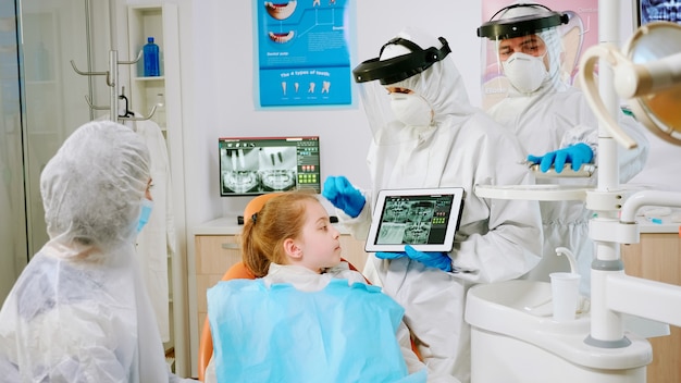 Stomatolog w sprzęcie ochronnym pokazujący na tablecie prześwietlenie dentystyczne przeglądające go z matką pacjenta. Zespół medyczny noszący maskę na twarz, rękawiczki, wyjaśniający radiografię za pomocą wyświetlacza notebooka;