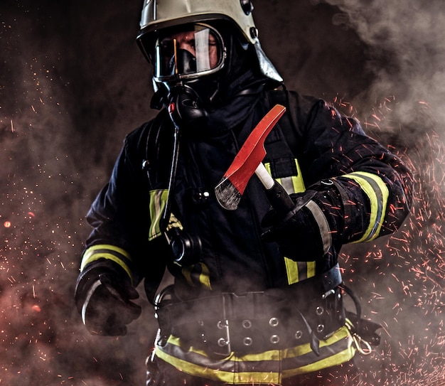 Strażak ubrany w mundur i maskę tlenową trzyma czerwoną siekierę stojącą w ognistych iskrach i dymie na ciemnym tle.