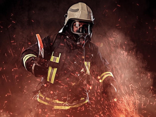 Bezpłatne zdjęcie strażak ubrany w mundur i maskę tlenową trzyma czerwoną siekierę stojącą w ognistych iskrach i dymie na ciemnym tle.