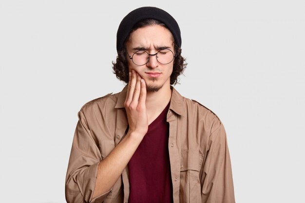 stresujący młodzieniec z małą brodą trzyma rękę na policzku, cierpi na ból zęba, trzyma oczy zamknięte, ubrany w stylowe ubrania, duże okrągłe okulary, modele na białej ścianie.